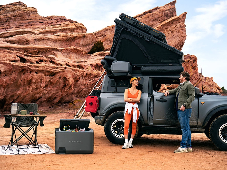Чоловік і жінка стоять, спираючись на великий автомобіль з тентом і кондиціонером на даху. Поруч з ним — холодильник EcoFlow.