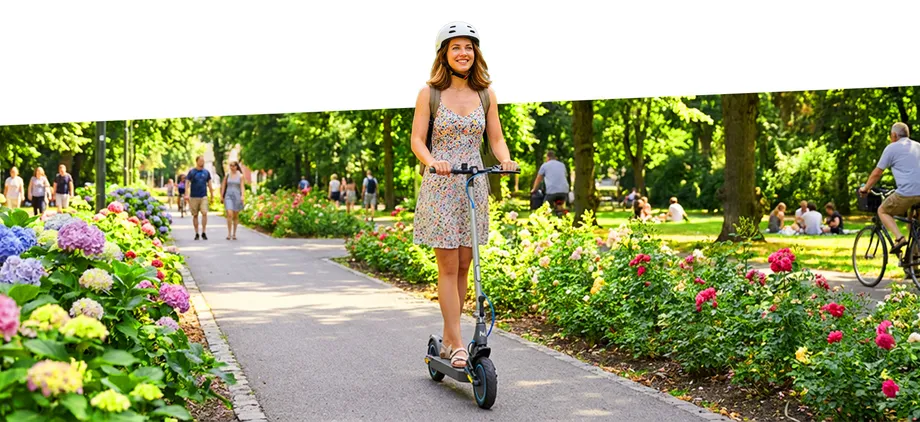 Дівчина в шоломі Motus їде на самокаті Scooty 10 Elite по парковій алеї серед людей, що йдуть.