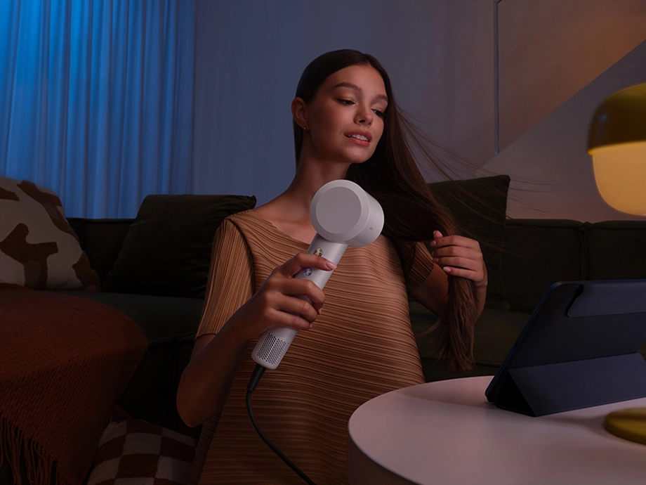 Жінка в бежевому светрі використовує білий фен, сидячи на дивані при вечірньому освітленні