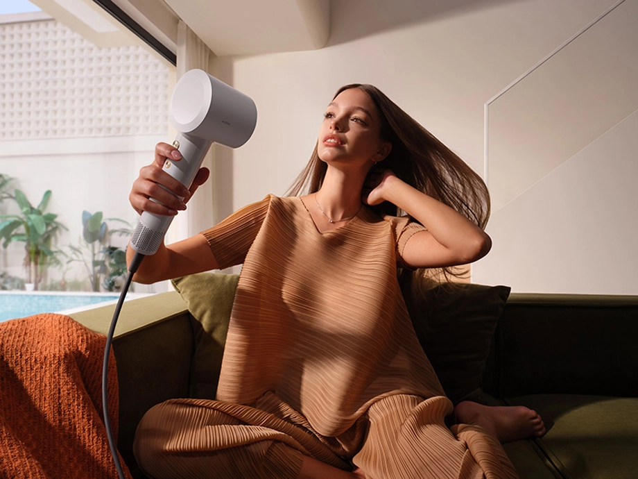 Жінка в бежевому светрі використовує білий фен, сидячи на дивані з видом на басейн