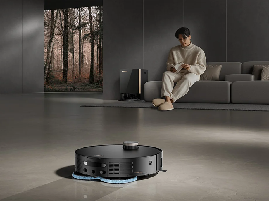 Чорний робот-пилосос миє підлогу в сучасній вітальні, поки чоловік сидить на дивані та читає книгу.