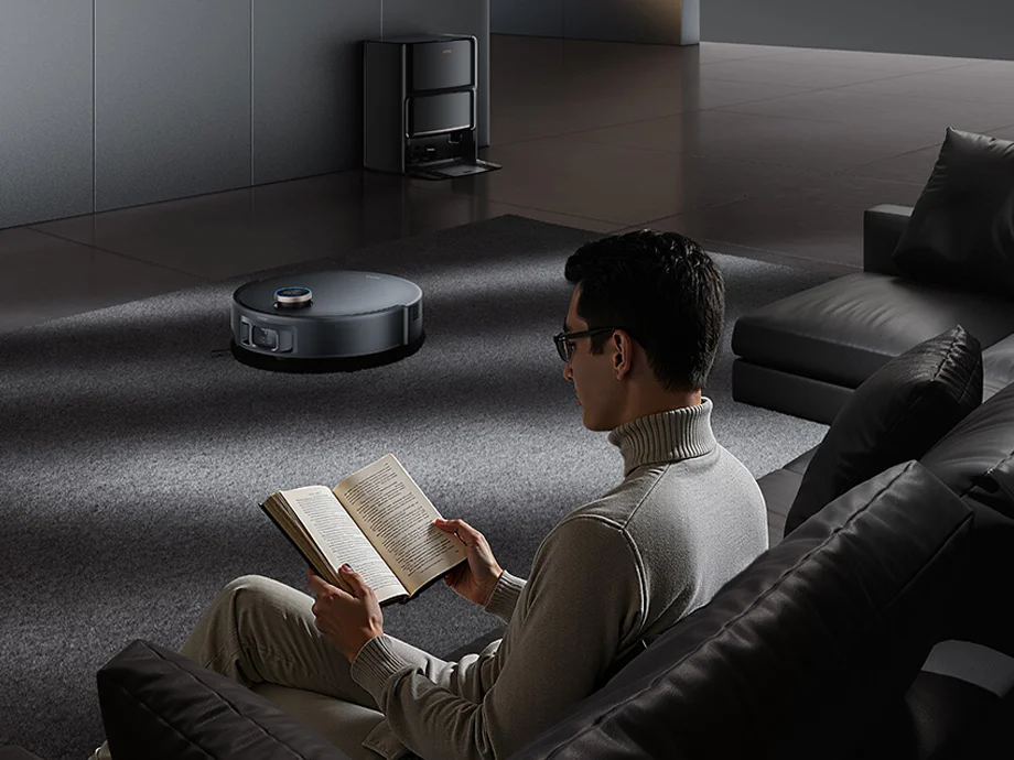 Чоловік сидить на дивані та читає книгу, поки робот-прибиральник працює на килимі на задньому плані.