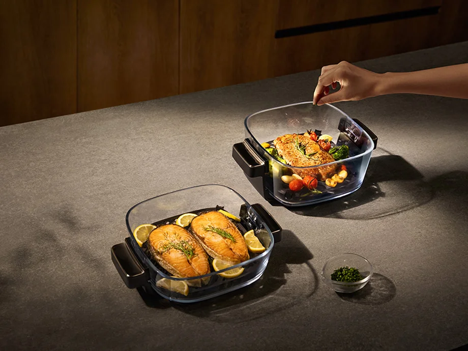 Дві прозорі форми з смаженим лососем та овочами, одну з них посипають спеціями руки людини.