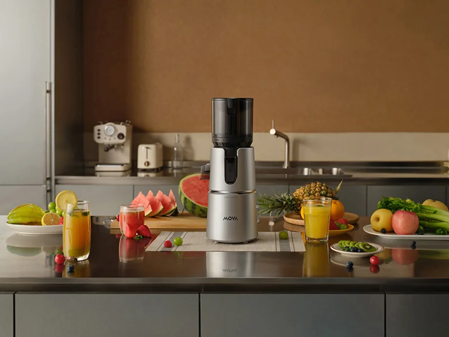 Повільна соковижималка стоїть на кухонній стільниці, оточена фруктами, овочами та різнокольоровими соками.