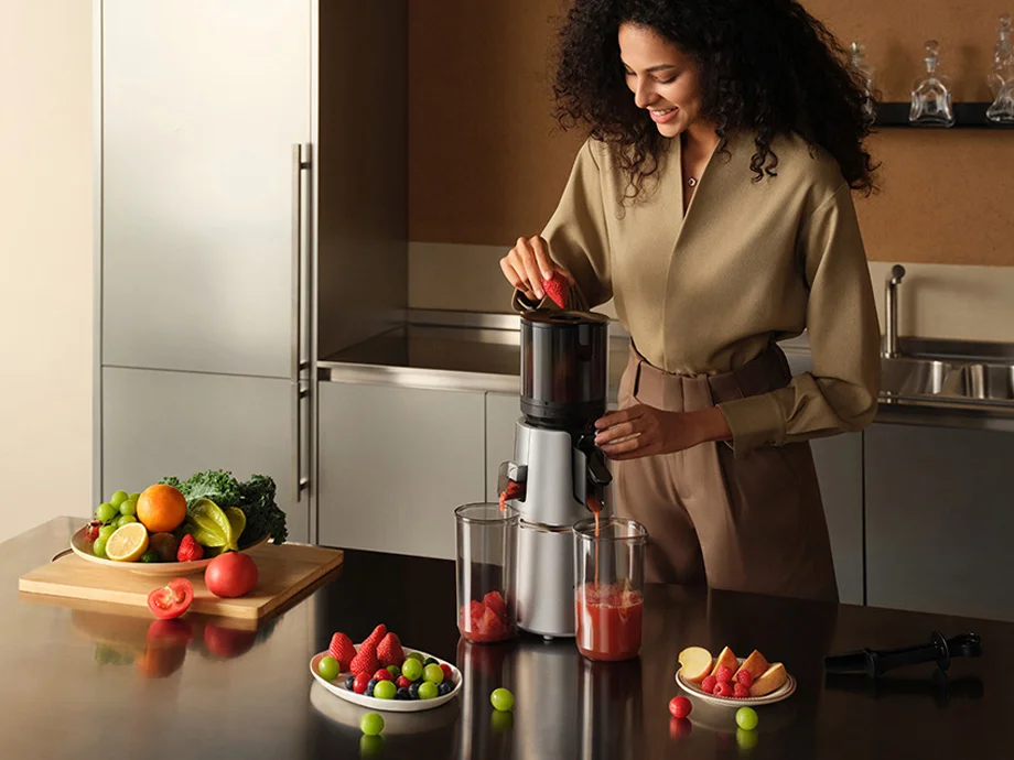 Жінка кладе скибочки кавуна в повільну соковижималку на кухонній стільниці поруч із фруктами та овочами.