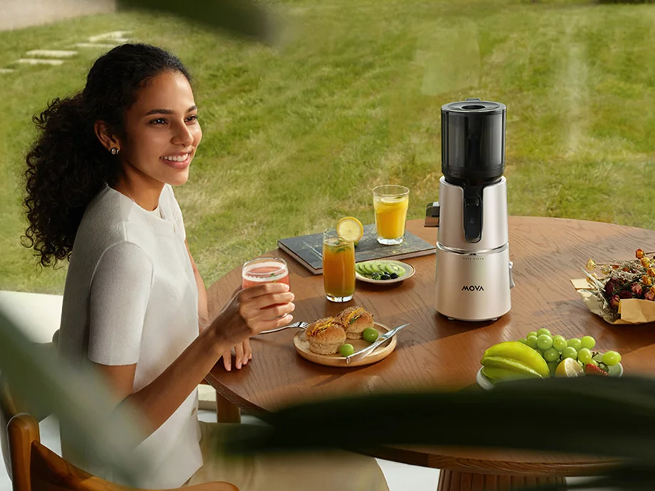 Жінка сидить за круглим столом з їжею та соками, тримаючи склянку соку поруч із повільною соковижималкою.