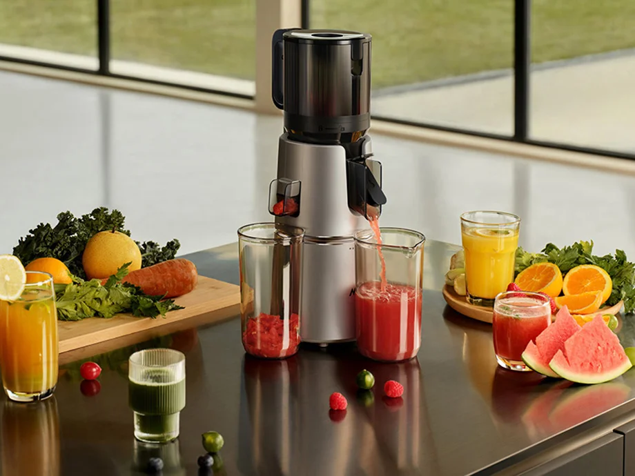 Повільна соковижималка наливає червоний сік у склянку, а навколо нього — фрукти, овочі та кольорові соки.