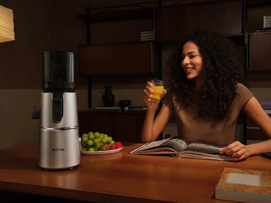 Жінка сидить за столом з книгою та склянкою соку, поруч із якою стоїть соковижималка та тарілка з фруктами.