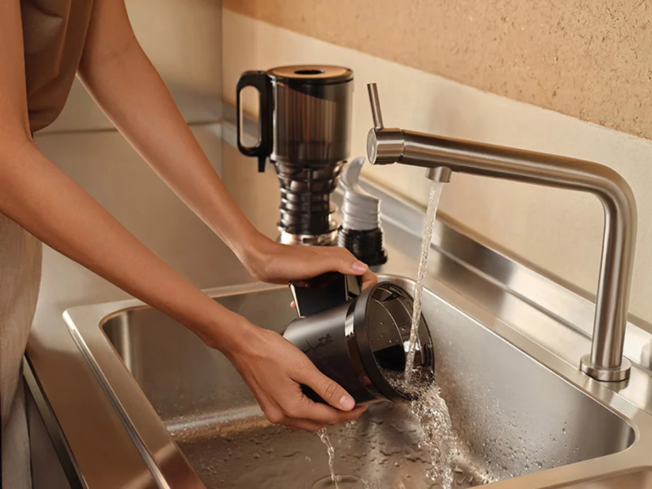 Жінка промиває елемент повільної соковижималки під проточною водою в кухонній раковині.