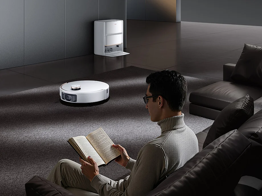 Чоловік сидить на дивані та читає книгу, поки робот-прибиральник працює на килимі на задньому плані.