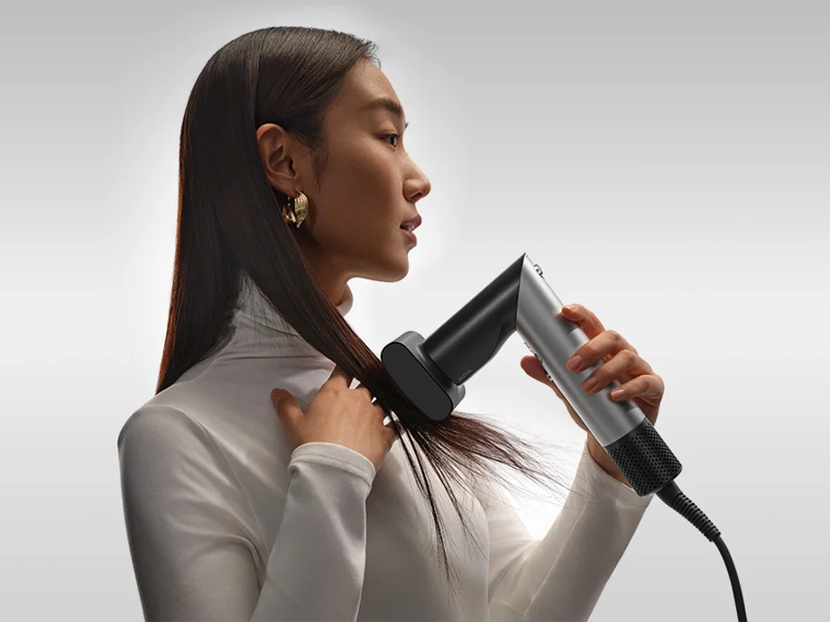 Жінка укладає своє пряме волосся феном із широким носом на сірому фоні