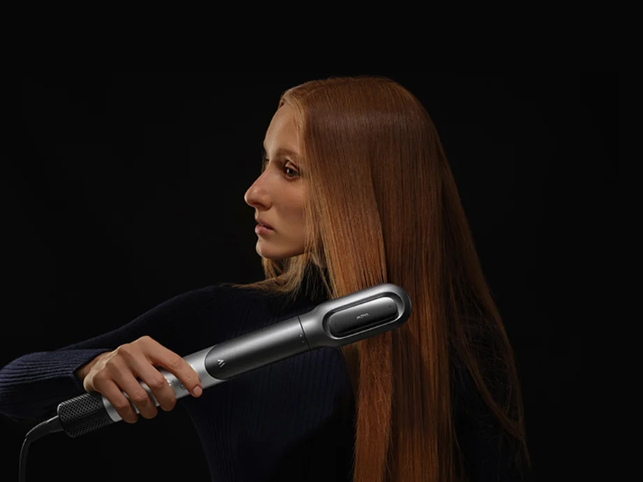 Жінка в темному светрі з високим коміром використовує пристрій, щоб пригладити своє довге руде волосся.