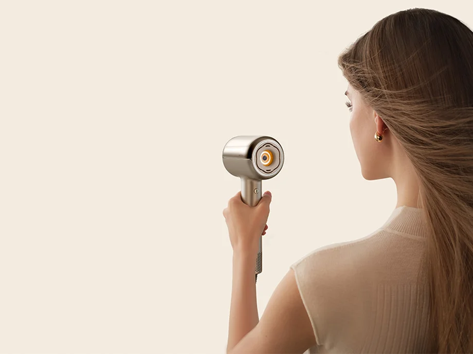 Жінка, зображена ззаду, тримає фен, який продуває повітрям її довге волосся.