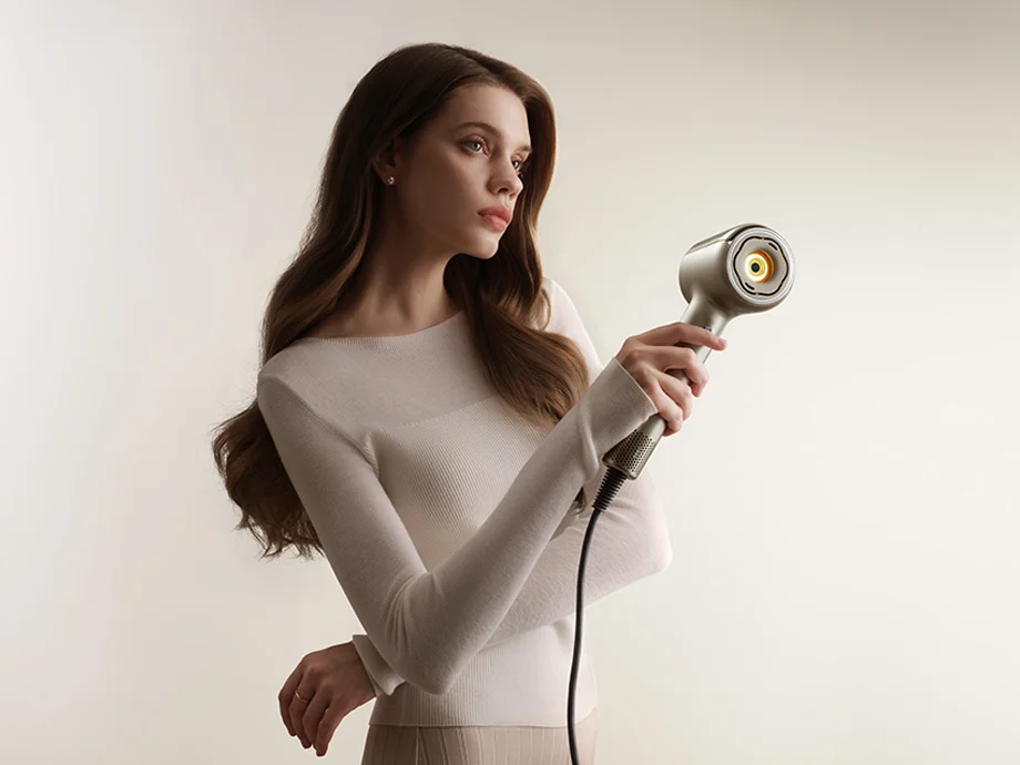 Жінка з довгим волоссям дивиться на фен, тримаючи його в руці.