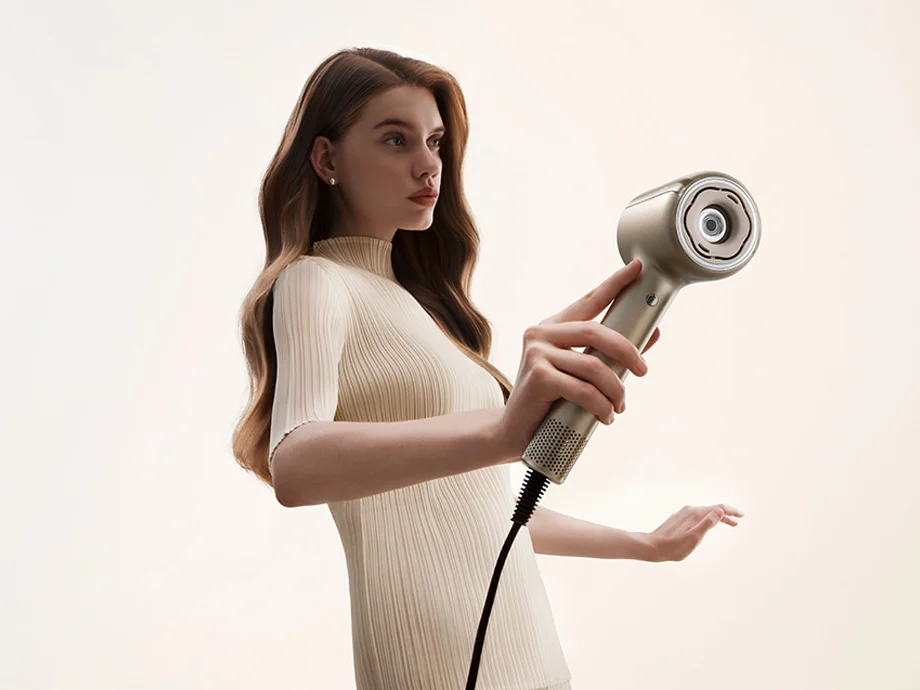 Жінка у легкій сукні тримає фен для волосся та дивиться вбік.
