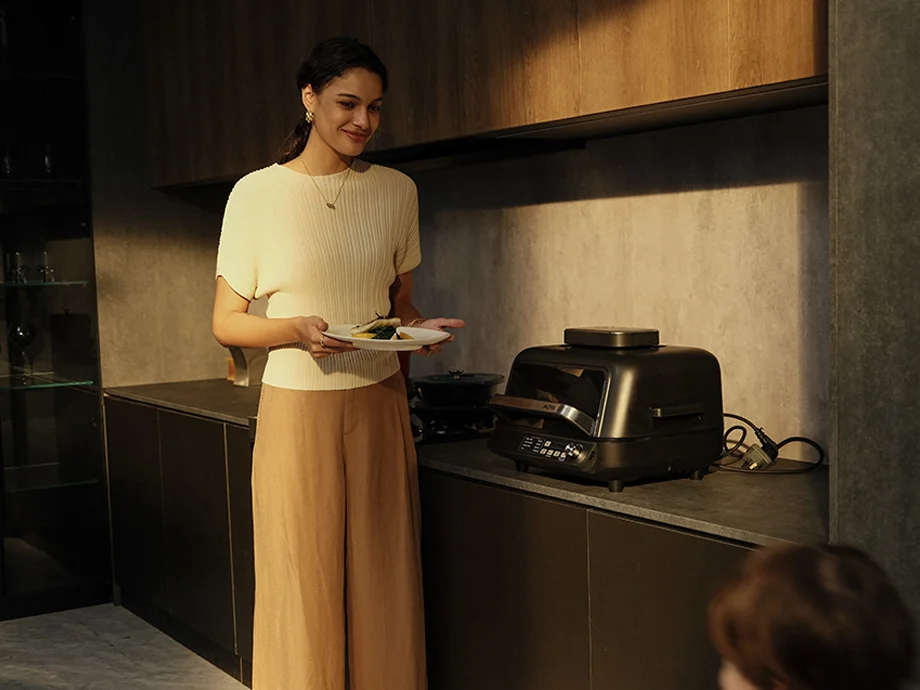 Жінка у білій футболці тримає тарілку з їжею, ліворуч від неї на кухонній стільниці знаходиться гриль.