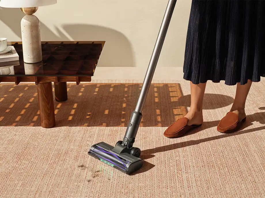 An upright vacuum cleaner collecting dirt from a light-colored carpet next to a coffee table.