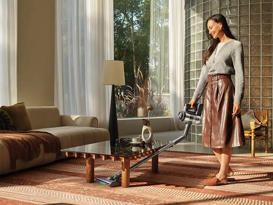 A woman cleans the floor under a low coffee table with an upright vacuum cleaner.