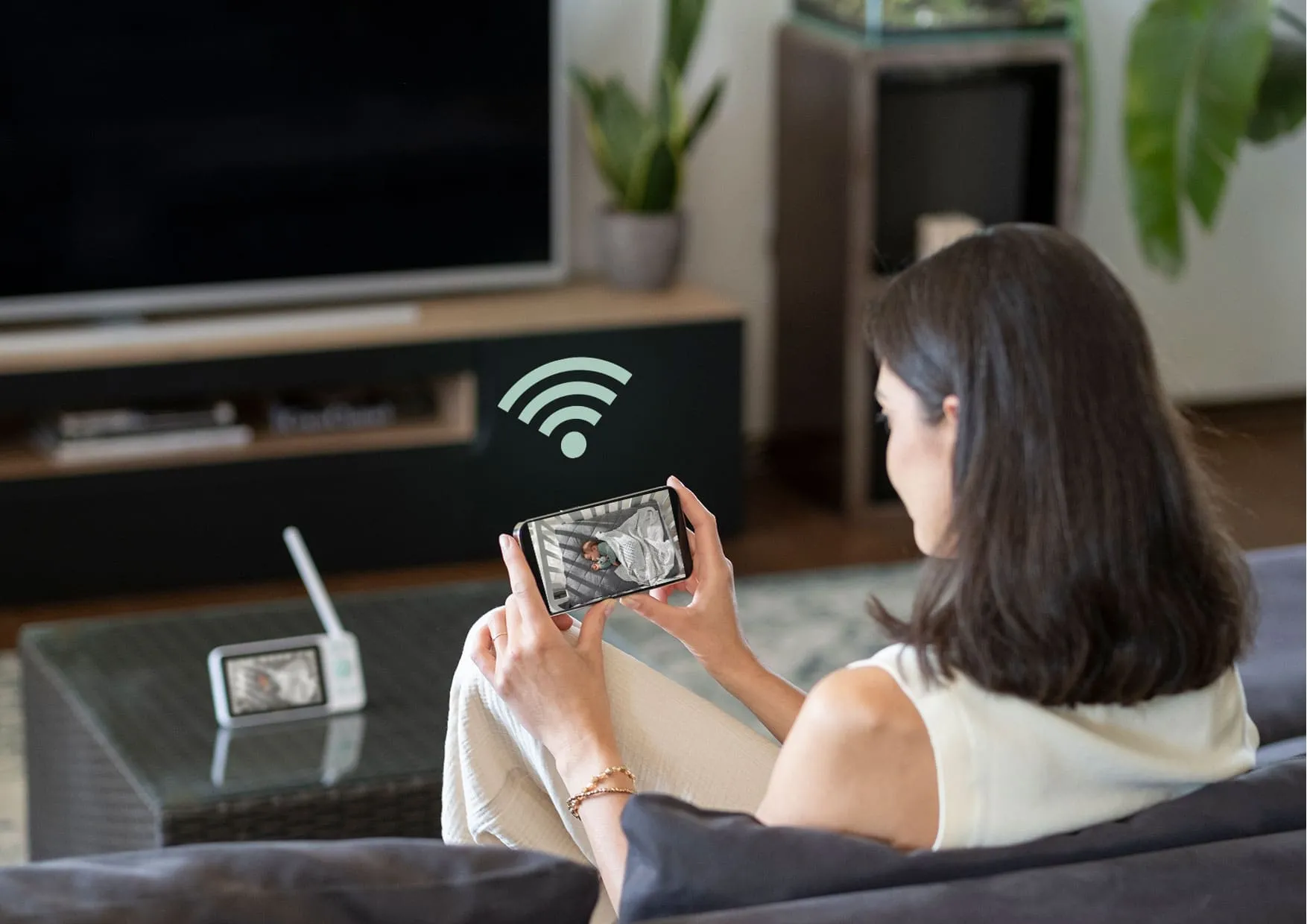 Жінка у вітальні спостерігає за сплячою дитиною на своєму смартфоні, на задньому плані — приймач Neno Ivo.