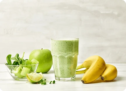 Густий зелений напій у склянці на кухонній стільниці, оточений бананами, яблуками, селерою та лаймами.