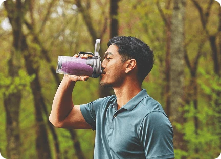 Чоловік у лісі п'є густий фіолетовий напій прямо з блендера Ninja Blast Max BC251EU через кришку з носиком.