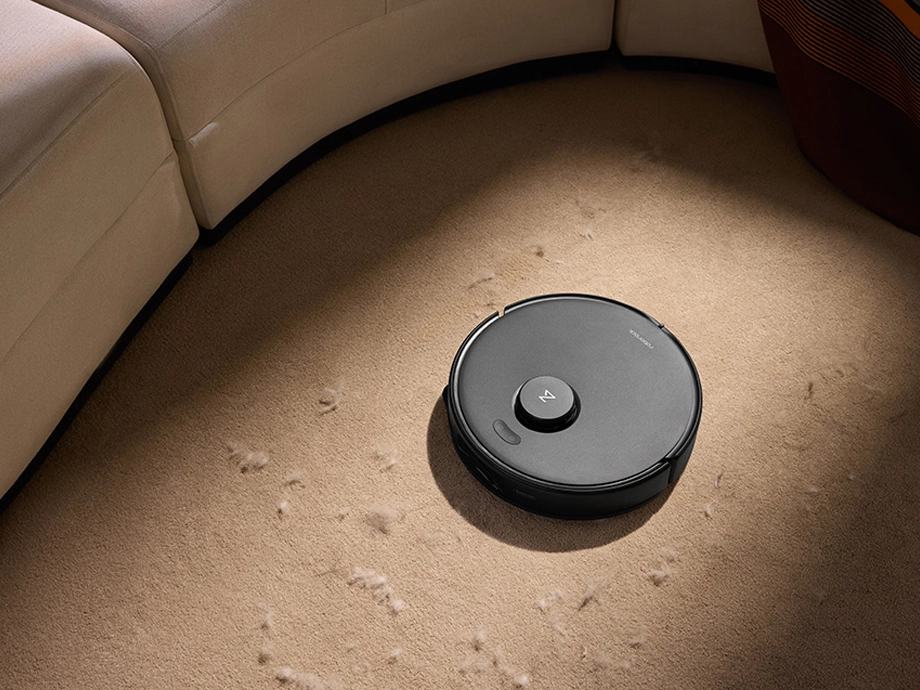 Робот-пылесос пылесосит бежевый пушистый ковер рядом с круглым диваном.