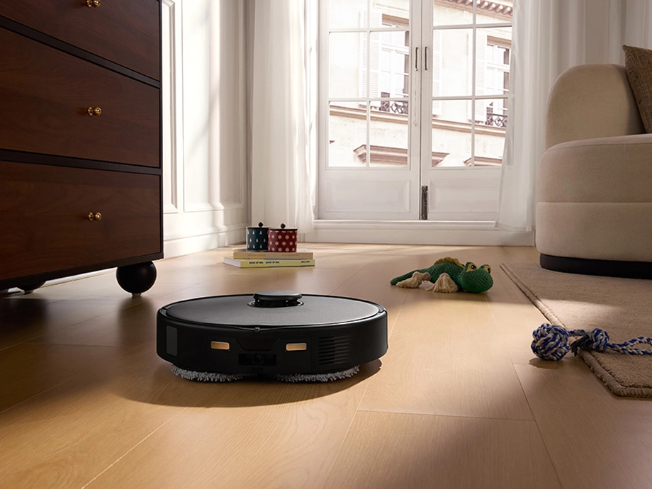 Робот-уборщик работает на деревянном полу рядом с игрушками и книгами у окна.
