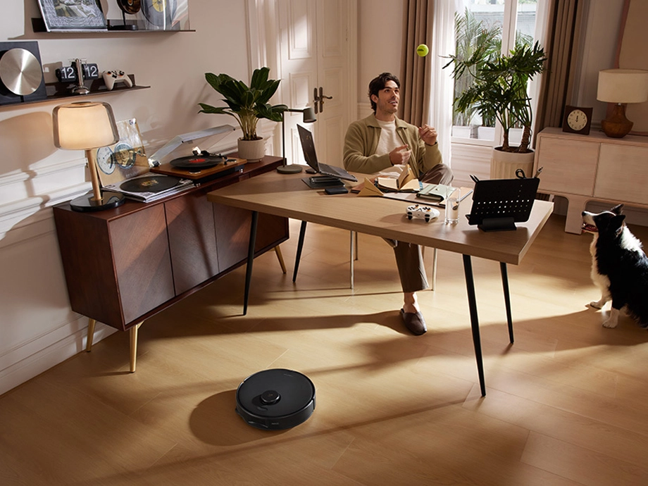 Мужчина сидит за столом и бросает мяч, в то время как робот-пылесос убирает пол.