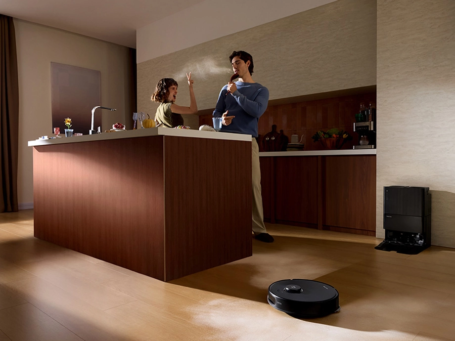 Мужчина и женщина стоят у кухонного острова, робот-пылесос работает на полу, а рядом с ним находится док-станция.