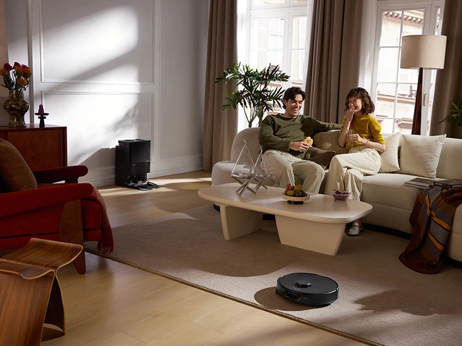 В гостиной мужчина и женщина сидят на диване, разговаривают и едят, в то время как робот пылесосит ковер.