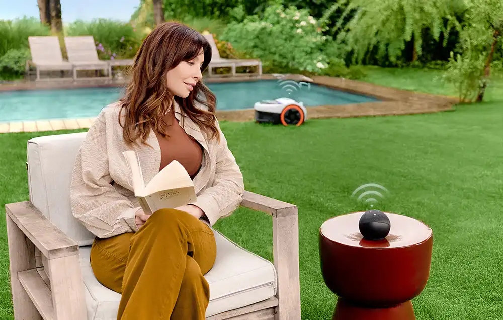 жінка сидить на стільці в саду, на задньому плані робот косить газон біля басейну