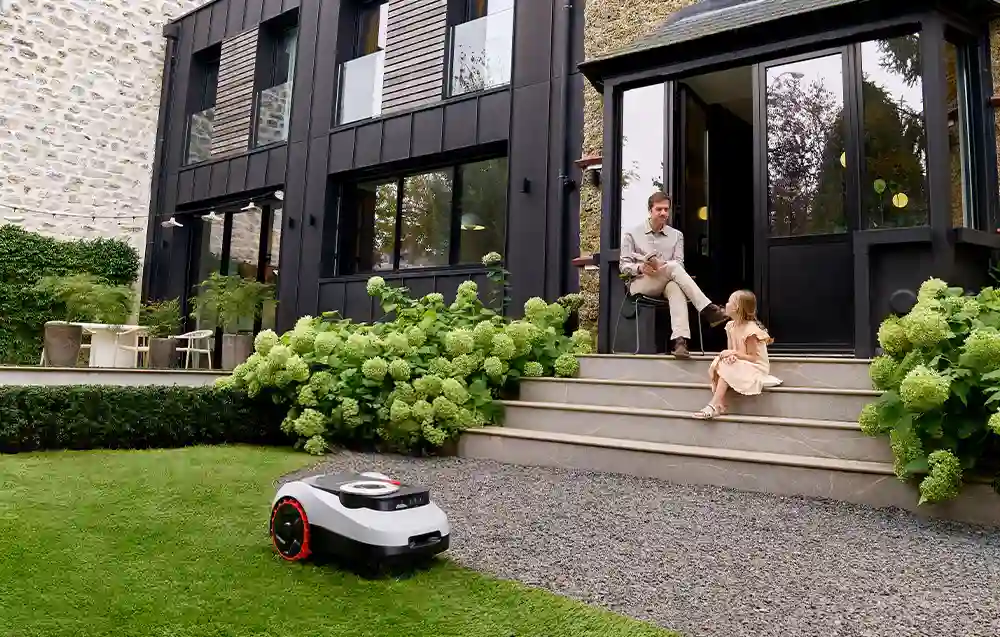 робот косить траву перед будинком, дівчина сидить на сходах