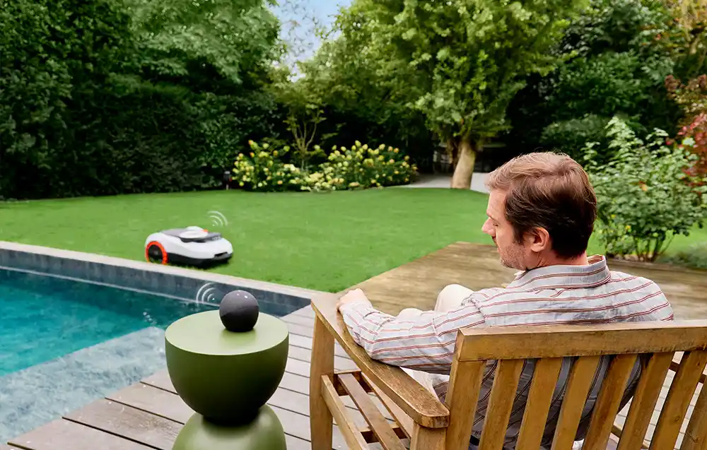 чоловік сидить на шезлонгу в саду, перед басейном, на задньому плані робот косить траву