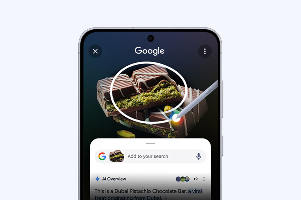 Na ekranie smartfona w wyszukiwarce Google pojawia się częściowo rozpakowany batonik czekoladowy Dubai Pistachio, oznaczony białym kółkiem i rysikiem. Poniżej znajdują się opcje wyszukiwania, opis określający go jako viralowy przysmak z Dubaju oraz fraza „Zakreśl, aby wyszukać w Google”.